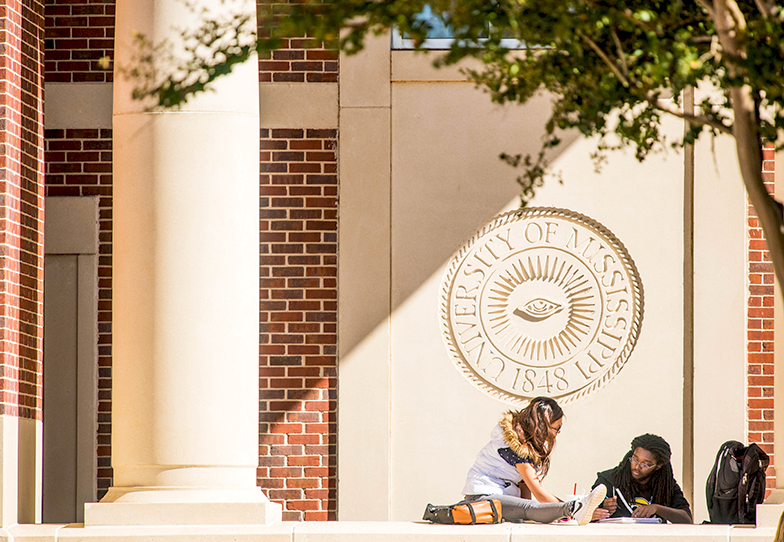 Two students study in front of the University of Mississippi crest engraved in the J.D. Williams Library wall outside.