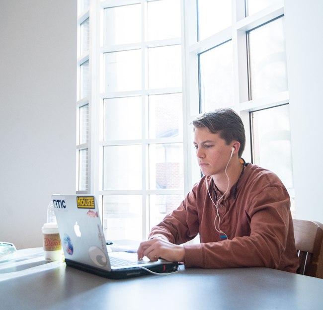 A student working on a laptop at a library table next to large windows.