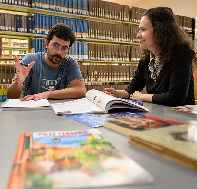 A student and mentor talking at a library table with open books spread out in front of them.
