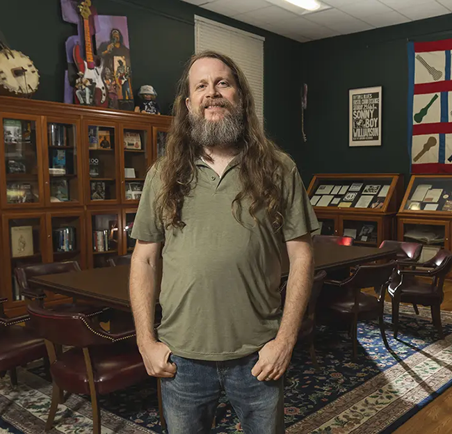 A bearded man with long hair stands in a library room lined with display cases full of vinyl records, photographs and other memorabilia. Posters and other artwork line frame the display cases.